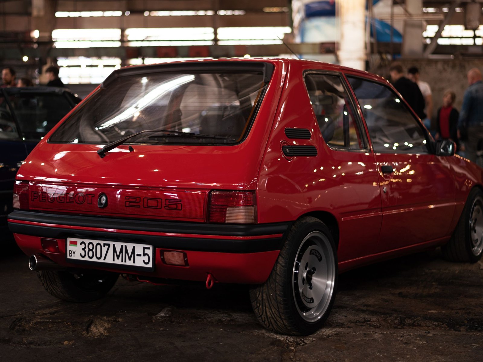 Peugeot 205 GTI rouge de trois quarts avant sur une route de campagne ensoleillée